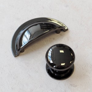 May include: Two black cabinet knobs, one is a round knob and the other is a curved, half-moon shaped knob.