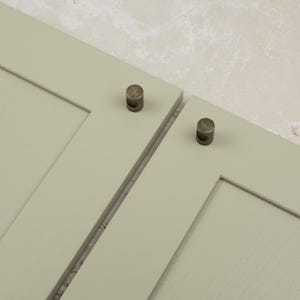 May include: Two sage green cabinet doors featuring a Shaker-style frame design. Each door is fitted with a small, cylindrical, antique brass-toned metal knob that has a recessed circular detail on the side, providing a functional grip.