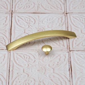 May include: A set of two brass-coloured cabinet hardware pieces. One is a curved handle, and the other is a small, round knob. The hardware has a brushed finish and is designed for use on drawers or cabinets. The hardware is shown against a patterned background.