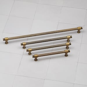 May include: Four antique brass cabinet pulls of varying lengths. Each pull features a textured, cylindrical bar with decorative end pieces. The pulls are arranged diagonally on a white tiled surface, showcasing their different sizes and designs.