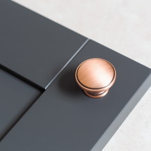 May include: A copper cabinet knob with a brushed finish. The knob is mounted on a dark gray cabinet door.