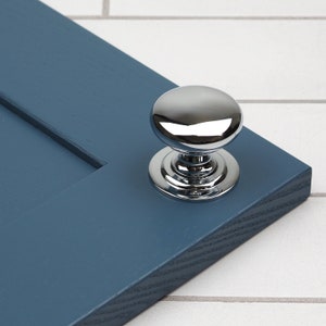 以下が含まれることがあります： 丸いドーム型のトップと滑らかで反射する表面を持つ、磨かれたクロム製のキャビネットノブ。ノブは青い塗装のキャビネットドアに取り付けられています。