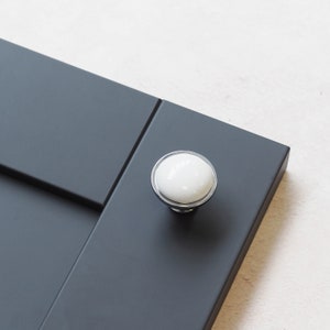 May include: A white ceramic knob with a silver metal base. The knob is mounted on a dark grey cabinet door.