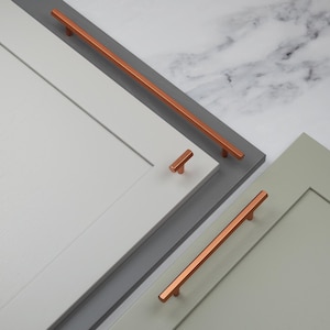 Pode incluir: Close-up de portas de armário com ferragens cor de cobre. As ferragens incluem puxadores longos e botões menores em forma de T. As portas são pintadas em tons de cinza e verde, com um fundo de mármore.