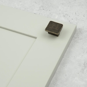 May include: A square, dark bronze-toned cabinet knob with a weathered, textured finish attached to a light sage green cabinet door panel. The knob features a flat top surface with slightly rounded edges and a tapered base connecting to the panel.