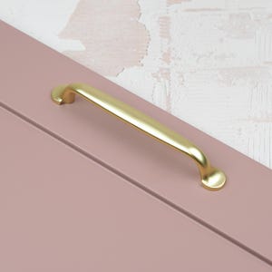 May include: A gold-coloured cabinet pull handle with a curved design is mounted on a light pink cabinet door. The handle has a smooth, polished finish and is positioned diagonally across the door. The background features a white and beige textured wall.