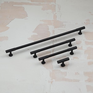 Può includere: Quattro maniglie nere per mobili di diverse lunghezze sono disposte su una superficie strutturata bianca e beige. Ogni maniglia ha una barra cilindrica con scanalature verticali e una base circolare. La più lunga misura circa 30 cm.