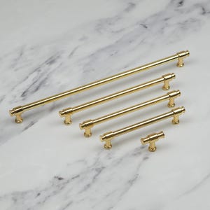 Op de afbeelding: Een verzameling goudkleurige kastgrepen in verschillende maten. De grepen hebben een cilindrisch staafontwerp met afgeronde eindkappen. De hardware is gerangschikt op een wit en grijs marmeren oppervlak.