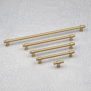 Pode incluir: Uma coleção de cinco puxadores dourados para armários em vários tamanhos. Os puxadores têm um design de barra cilíndrica com extremidades arredondadas e estão dispostos sobre uma superfície texturizada esbranquiçada. Ferragens para móveis e armários.