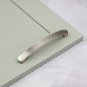 Peut inclure: Une poignée de porte de placard en nickel brossé avec un design incurvé. La poignée est montée sur une porte de placard vert clair.
