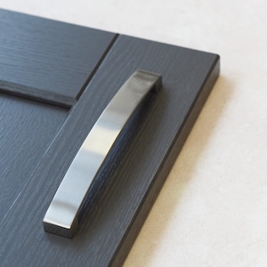 Puede incluir: Un tirador de armario de níquel cepillado está montado en una puerta de armario gris oscuro. El tirador tiene un diseño curvo y una base rectangular. La puerta del armario tiene una textura de veta de madera.