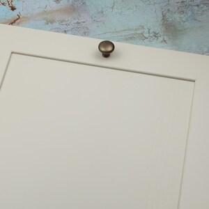 Peut inclure: Gros plan sur une section d'une porte d'armoire cr&egrave;me avec un bouton de couleur bronze. Le bouton est rond et l&eacute;g&egrave;rement bomb&eacute;. L'arri&egrave;re-plan est un mur textur&eacute; avec des nuances de bleu, de marron et de blanc.