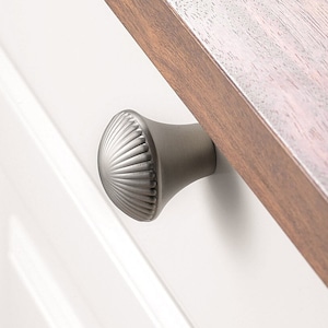 以下が含まれることがあります： 白いキャビネットのドアに取り付けられた、フルーテッドの貝殻の形をした銀色のキャビネットノブ。