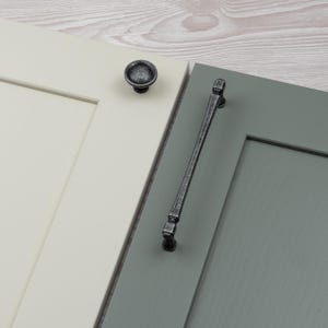 Pode incluir: Close-up de duas portas de armário com ferragens. Uma porta é creme com um puxador redondo de metal escuro. A outra porta é cinza-esverdeada com uma puxador longo de metal escuro. As ferragens têm um acabamento antigo.