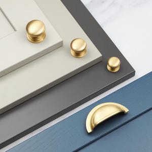 Könnte beinhalten: Vier goldene Schrankknöpfe und ein goldener Schrankzug auf verschiedenen farbigen Oberflächen. Die Knöpfe sind rund und haben eine glatte Oberfläche. Der Zug ist halbkreisförmig.