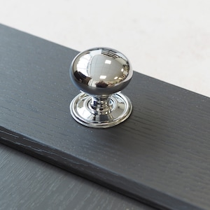 May include: A chrome knob with a round, polished surface. The knob is mounted on a dark gray wooden surface.
