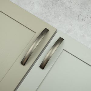 May include: Two cabinet doors featuring a flat-panel design with a smooth finish. One door is a warm beige, and the other is a light gray. Each door is fitted with a curved, dark metallic handle with a brushed, weathered texture.