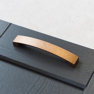 May include: A brushed gold cabinet pull with a curved design. The pull is mounted on a dark wood cabinet door.