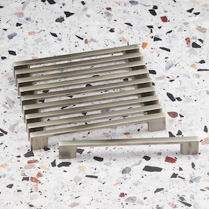 May include: A set of ten silver cabinet pulls with a rectangular shape and a brushed finish. The pulls are arranged in a stack, with one pull lying horizontally in front of the stack.