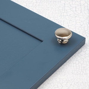 Pode incluir: Uma maçaneta de armário de níquel polido com uma parte superior redonda ligeiramente abobadada. A maçaneta está montada em uma porta de armário pintada de azul.