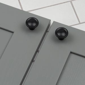 Può includere: Due pomelli per armadietti neri con un design rotondo leggermente rialzato sono montati su ante di armadietti grigie.