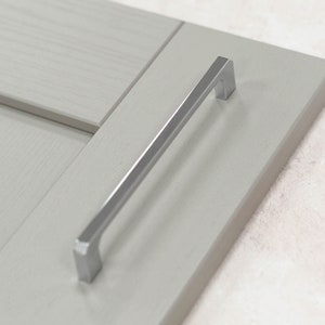 May include: A brushed nickel cabinet pull with a rectangular shape and a slightly curved design. The pull is mounted on a light gray cabinet door.