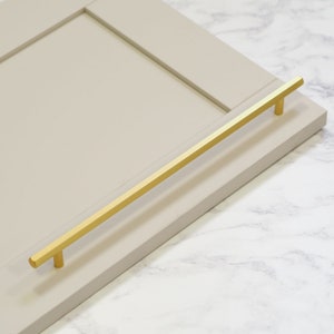 May include: A gold metal cabinet pull with a rectangular shape. The pull is mounted on a light gray cabinet door.