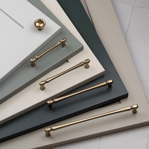 May include: A selection of cabinet handles and a knob in a brass finish, presented on painted wooden panels in various shades of grey and blue. The hardware features a classic, elegant design.