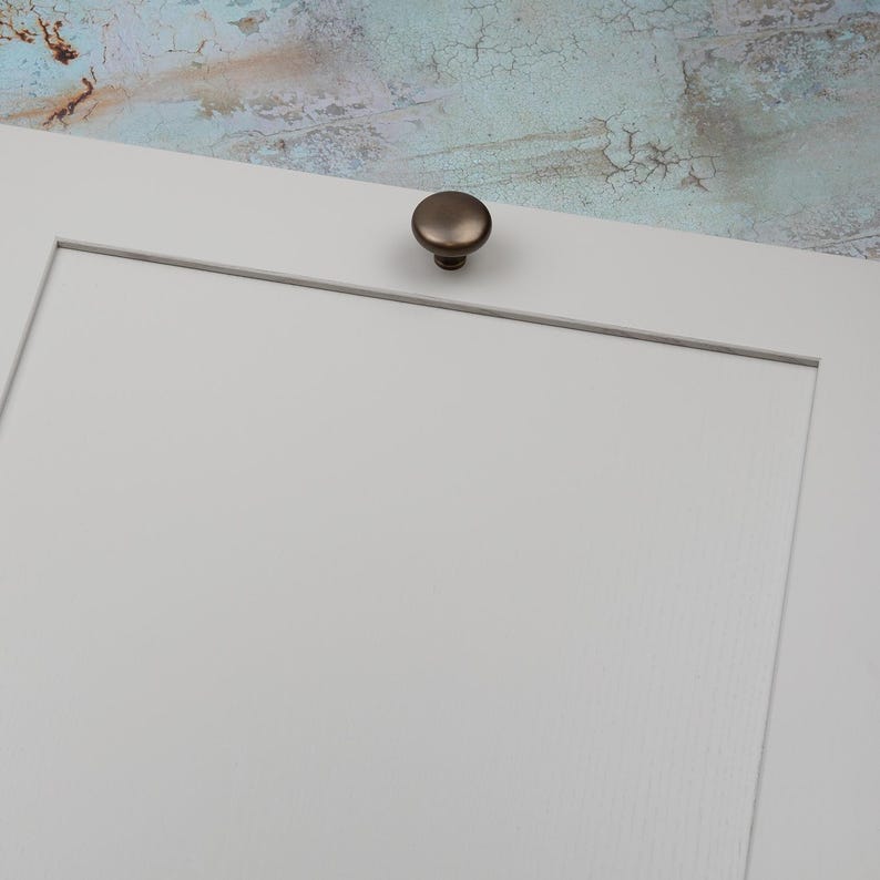 Peut inclure: Gros plan sur une porte d'armoire blanche avec un bouton de couleur bronze. Le bouton est rond et l&eacute;g&egrave;rement bomb&eacute;. L'arri&egrave;re-plan est une surface textur&eacute;e et multicolore.