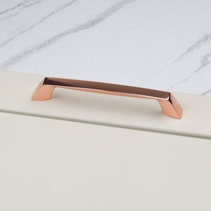 Könnte beinhalten: Ein roségoldener Schrankgriff mit geometrischem Design. Der Griff ist rechteckig mit abgewinkelten Enden und ist auf einer hellen Oberfläche montiert. Der Griff ist aus Metall und hat eine polierte Oberfläche.