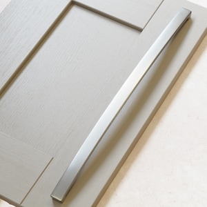 May include: A brushed nickel cabinet pull with a rectangular shape and rounded edges. The pull is mounted on a light gray cabinet door.