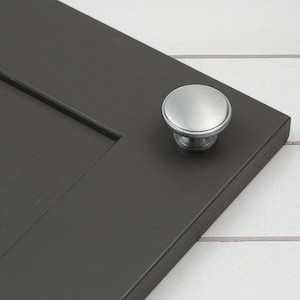 May include: A brushed nickel cabinet knob with a round, slightly raised top. The knob is mounted on a dark gray cabinet door.