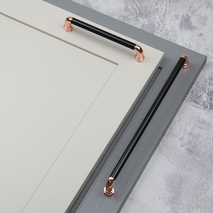 Op de afbeelding: Kast hardware met zwarte stanggrepen en koperkleurige accenten. De grepen zijn gemonteerd op een witte en grijze kastdeur. Modern, industrieel ontwerp.