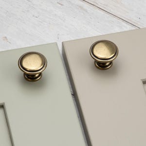 May include: Two round cabinet knobs with an antique brass finish, each featuring a smooth, slightly domed surface and a stepped base. One knob is mounted on a light sage green panel, and the other is on a light beige panel, both showing hardware.