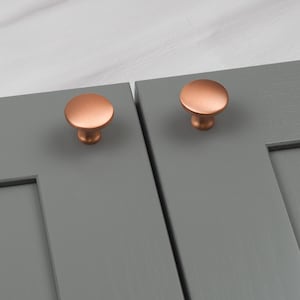 以下が含まれることがあります： グレーのキャビネットドアに取り付けられた2つの銅色のキャビネットノブ。ノブは丸みを帯びたキノコ型のデザインです。キャビネットドアは無地のグレーに塗装されています。