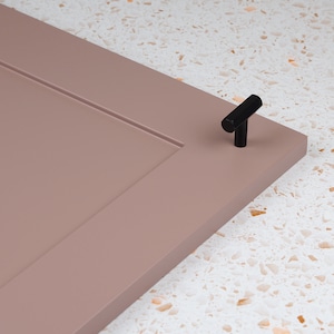 May include: A close-up of a dusty rose cabinet door with a black T-shaped cabinet knob.
