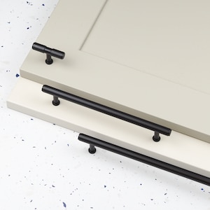 May include: Three black cabinet pulls, two are long and one is short and T-shaped. The pulls are shown on a white cabinet door.