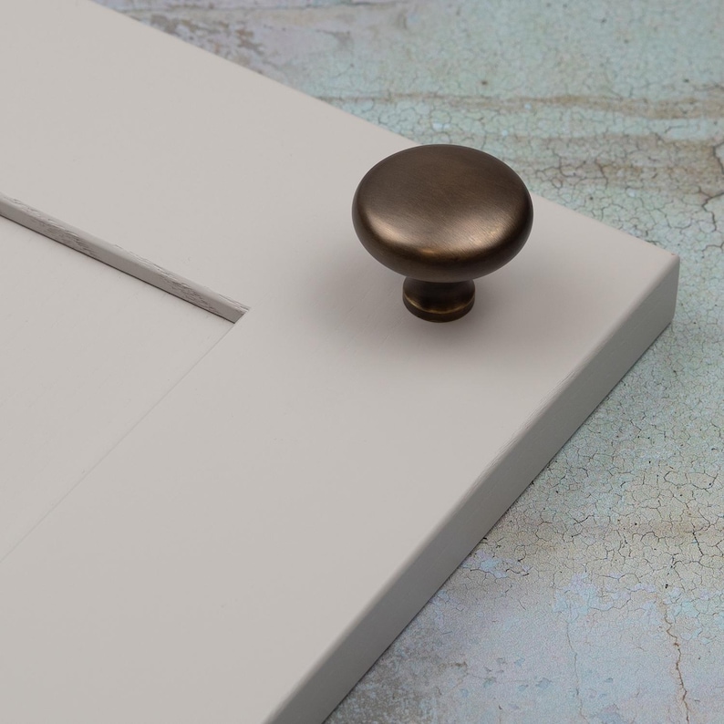 Peut inclure: Un bouton de meuble en bronze avec un dessus arrondi et une base cylindrique courte. Le bouton est mont&eacute; sur une section d'une porte d'armoire gris clair. L'arri&egrave;re-plan a un aspect mouchet&eacute; et vieilli.