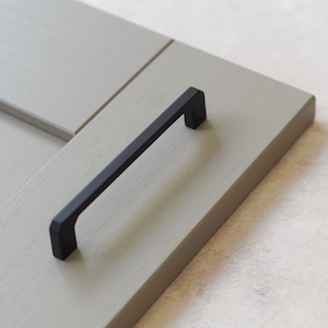 May include: Black cabinet door handle with a rectangular shape. The handle is mounted on a light gray cabinet door.