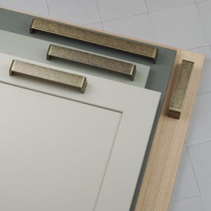 Peut inclure: Gros plan de plusieurs poignées de meubles en laiton antique de différentes tailles. Les poignées rectangulaires ont une surface légèrement texturée. Elles sont présentées sur une porte d'armoire gris clair et une surface en bois.