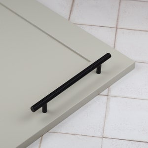 May include: Black cabinet hardware pull with a rectangular shape and rounded ends. The pull is mounted on a light gray cabinet door.