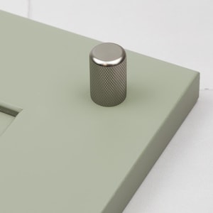 May include: A silver knob with a textured finish, mounted on a light green cabinet door.