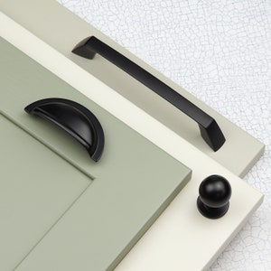 May include: A collection of black cabinet hardware, including a long bar pull, a cup pull, and a round knob. The hardware is displayed on painted wooden panels in shades of green, white, and cream.