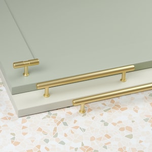May include: Three gold cabinet pulls, two are long and one is short. The pulls are on a light green and white surface.