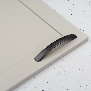 Pode incluir: Uma puxador de armário preto com acabamento escovado. O puxador está montado em uma porta de armário cinza claro.