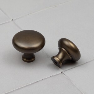 Peut inclure: Deux boutons de meuble de couleur bronze. Chaque bouton a un dessus arrondi et une base cylindrique courte. Un bouton est droit, l'autre est sur le c&ocirc;t&eacute;. Les boutons ont une finition lisse et mate. Fond gris clair.