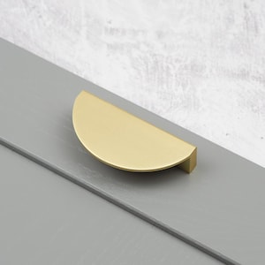 Peut inclure: Une poignée de meuble en forme de demi-lune avec une finition en laiton brossé. La quincaillerie est montée sur une surface en bois peinte en gris. La poignée est un élément de design moderne.
