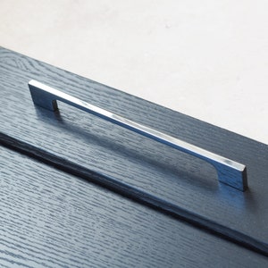 May include: A silver cabinet door handle with a rectangular shape and a slight curve. The handle is mounted on a dark wood cabinet door.