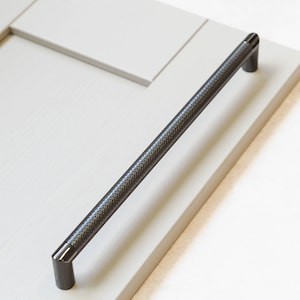 May include: A black metal cabinet pull with a textured finish. The pull is mounted on a white cabinet door.