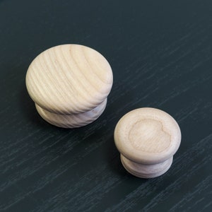 May include: Two wooden knobs, one larger than the other, are shown on a black surface. Both knobs are light brown and have a smooth, rounded shape.